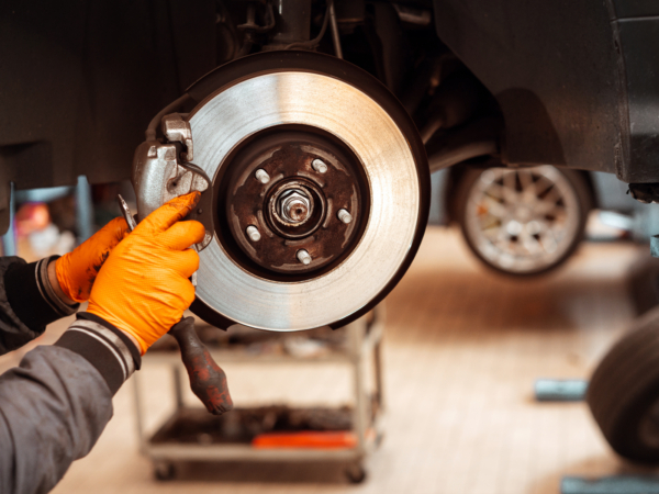 Anonymous mechanic in jacket and gloves while standing in workplace in automobile garage and replacing car wheel brake pad against blurred interior in daylight